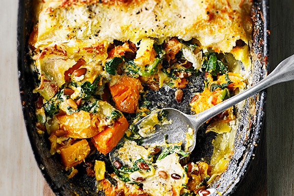 Butternut squash, spinach and mascarpone lasagne in a large oval black baking dish, placed on a wooden plank