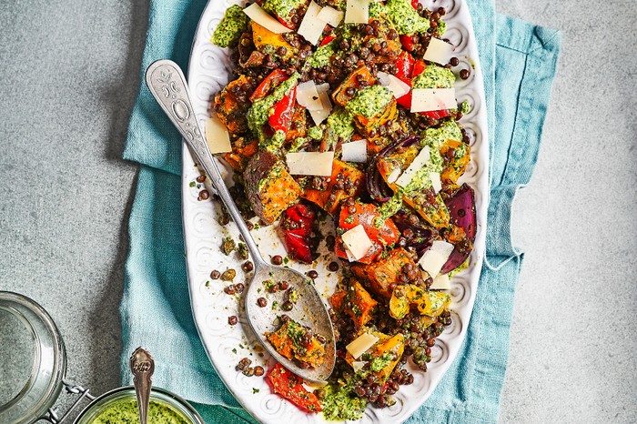 A grey background with a white platter, topped with roasted vegetables, grains and a green dressing