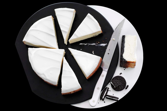 Black and white Oreo cheesecake cut into six pieces, next to a knife and placed on a black plate