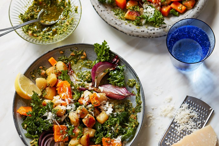A white marble background topped with two grey bowls, filled with golden gnocchi, green kale and cubes of squash