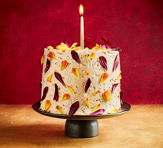 A birthday cake covered in purple, yellow and orange petals on a cakestand with a candle on the top