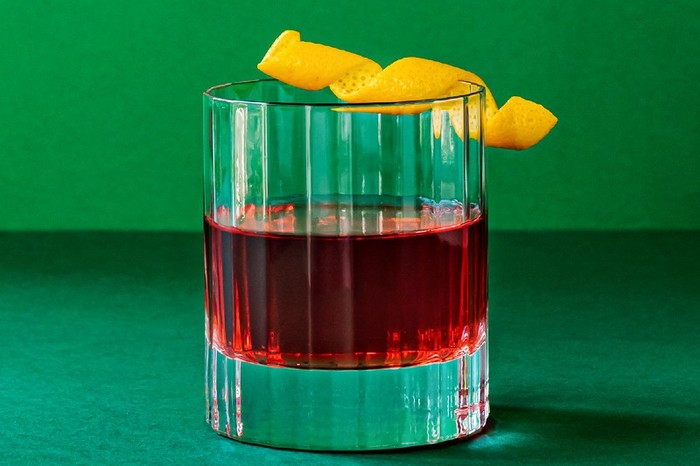 A dark red cocktail in a tumbler with a twist of orange peel on the rim