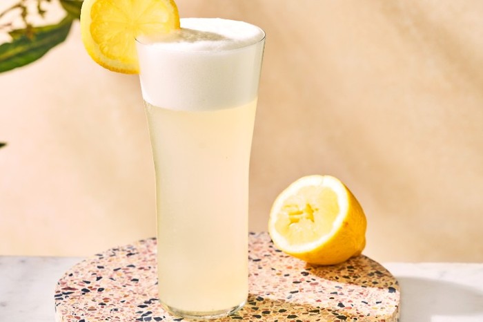 A tall glass filled with a light peach-coloured cocktail with a foamy top and garnished with a slice of lemon on a terrazzo board