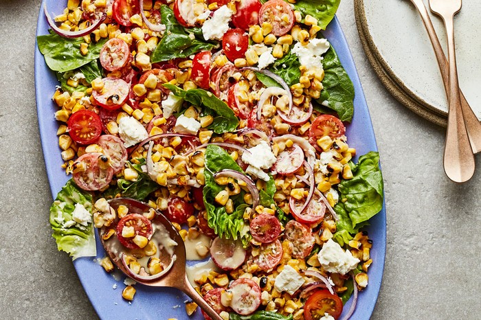 Salad of corn, lettuce, tomatoes, onions and feta on a blue plate next to a bowl of dressing