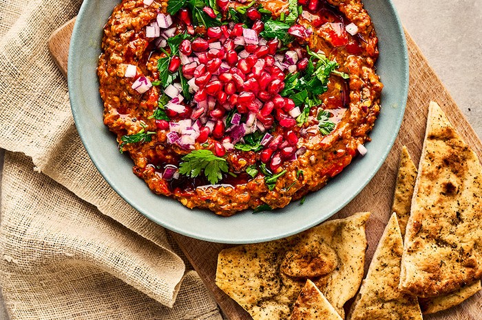 A bowl of muhammara topped with red onion, parsley and pomegranate seeds