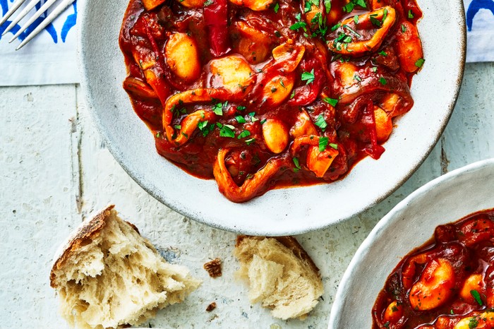 A bowl filled with a vibrant red squid, pepper, butter bean stew with torn bread on the side