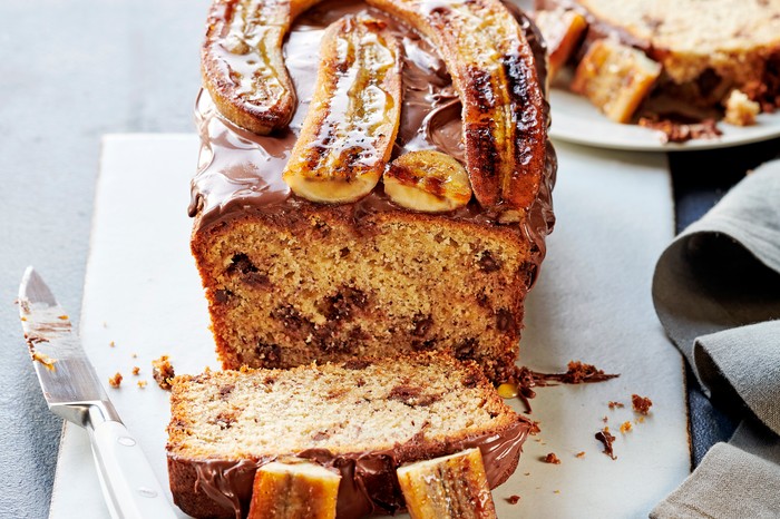 A banana and chocolate chip loaf cake on a white serving board, with a slice cut from the end