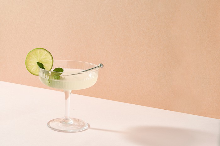 A small coupe glass filled with a lime cocktail with a wheel of lime and sprig of mint