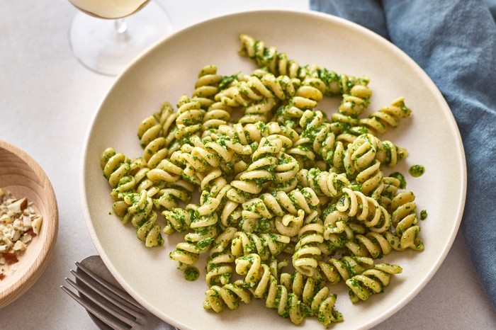 A bowl of fusilli pasta coated in green pesto next to a glass of wine and blue linen napkin