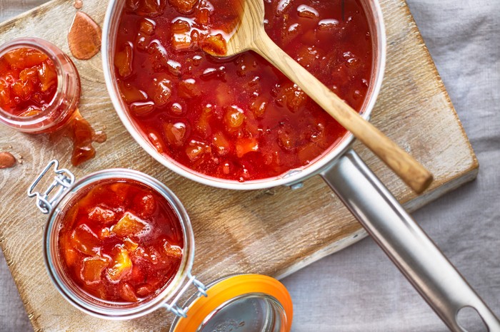 A silver saucepan filled with red jam and a wooden spoon next to a jar of jam on a wooden board