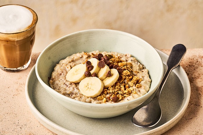 A bowl of porridge topped with banana, granola and chocolate chips, next to a coffee