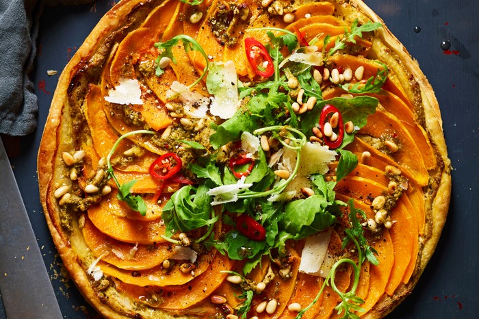 A round pastry tart topped with slices of orange pumpkin, green rocket leaves and pine nuts