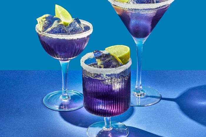 Three different shaped cocktail glasses filled with a purple drink, ice cubes and lime wedges on a blue background