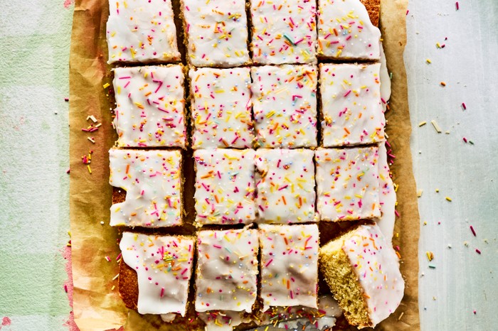 A school cake topped with icing and sprinkles cut into squares on a sheet of brown baking parchment