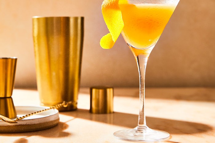 A sidecar cocktail with lemon peel garnish beside a bronze cocktail shaker