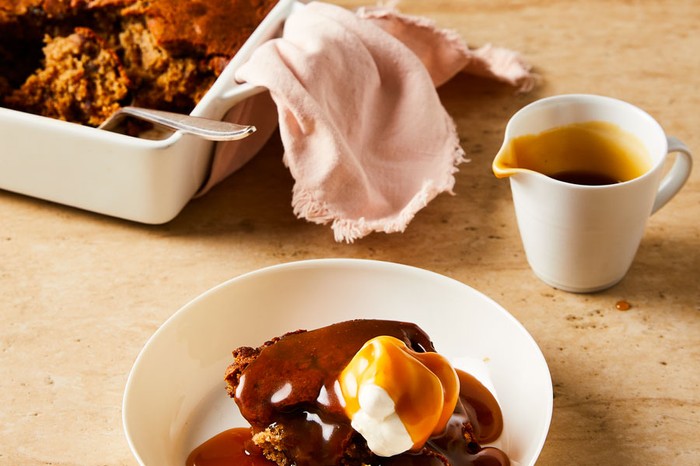 A bowl of sticky pudding topped with a dollop of cream, plus a jug of toffee sauce and a baking tray of sticky toffee pudding