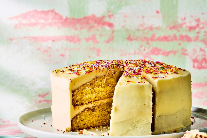 A vanilla sponge cake covered in icing and topped with sprinkles with a slice taken out, against a pink and green background