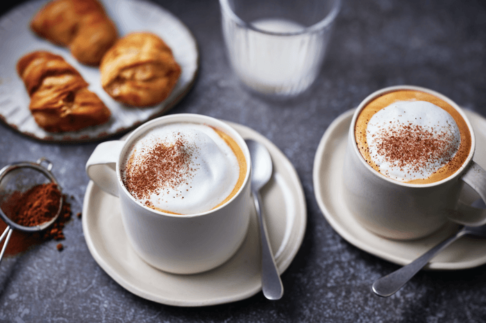 Two white cups of cappuccino with chococlate dusting and a plate of croissants
