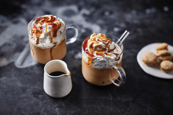 Two glass mugs of coffee topped with whipped cream and caramel sauce, with a small white jug of caramel