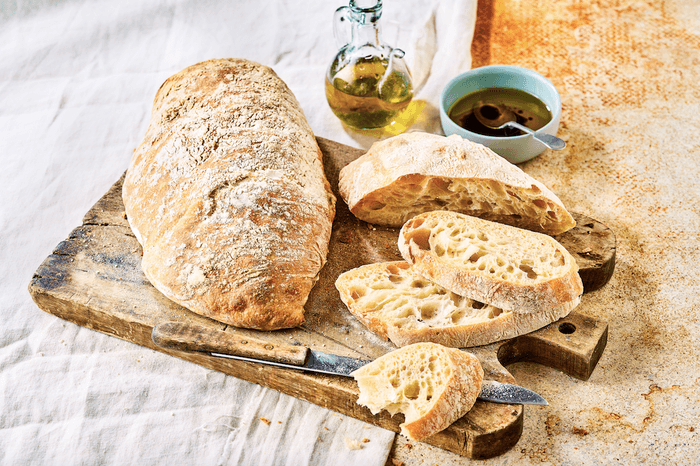 Two loaves of ciabatta on a wooden board with a bottle of oil and bowl of balsamic vinegar, one sliced into pieces